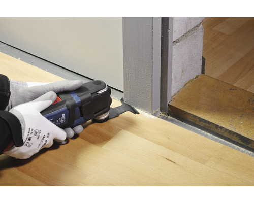 Osoba používa multifunkčný nástroj na rezanie drevenej podlahy pri dverách.