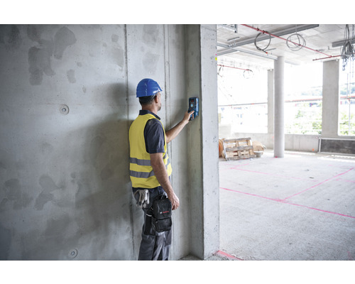Stavebný robotník s helmou a bezpečnostnou vestou používa diaľkomer na betónovom múre na stavenisku.