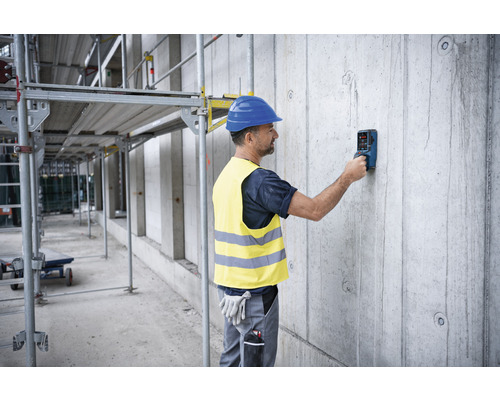 Muž s ochrannou prilbou a výstražnou vestou používa detekčné zariadenie na betónovej stene na stavenisku.