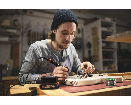 Muž spájkuje gitaru spájkovačkou na pracovnom stole.