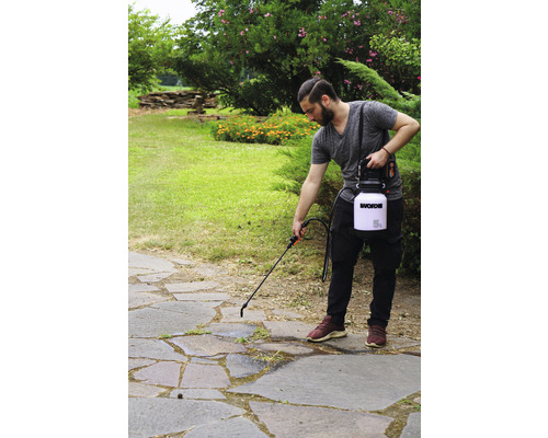 Osoba používa záhradný postrekovač na kamennej terase na ničenie buriny. V pozadí sú kríky a záhrada.