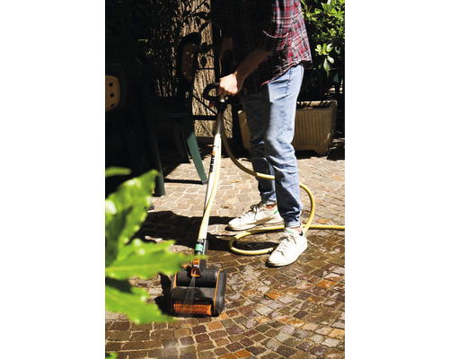 Muž čistí dlažobné kocky kefovým čističom a vodnou hadicou v záhrade.