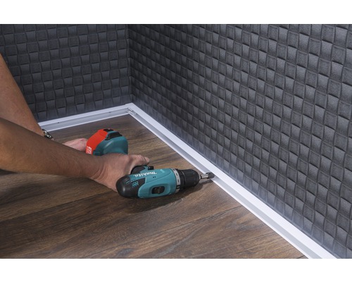 Remeselník montuje soklovú lištu pomocou akumulátorového skrutkovača v miestnosti s drevenou podlahou a štruktúrovanou stenou