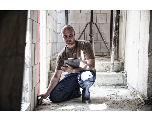 Muž používa laserový diaľkomer na stenu v hrubej stavbe.