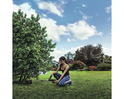 Žena pracuje s akumulátorovou kosačkou na trávu v záhrade na strome