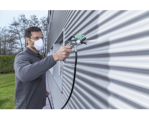 Muž s ochrannými okuliarmi a maskou strieka stenu farbou pomocou striekacej pištole