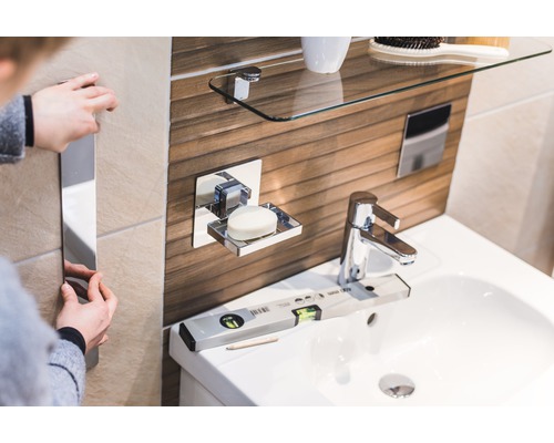 Svetlá kúpeľňa s umývadlom, mydelničkou, sklenenou policou a osobou, ktorá inštaluje zrkadlo