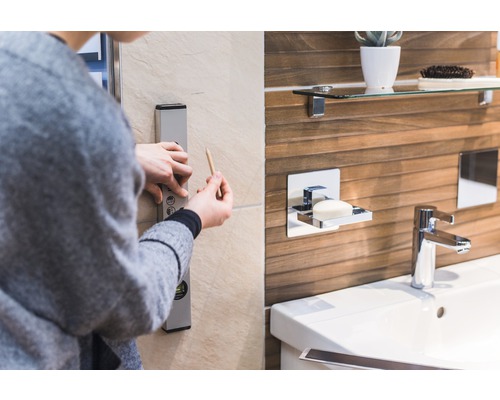 Osoba používa vodováhu a ceruzku v kúpeľni na označenie priamky na stene. V pozadí je vidieť umývadlo, vodovodný kohútik a dávkovač mydla.