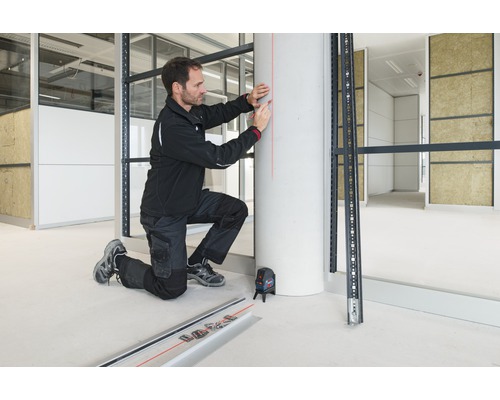 Remeselník s krížovým líniovým laserom pri montáži kovovej spodnej konštrukcie v interiéri.