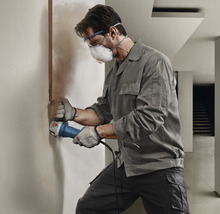 Remeselník pracuje s drážkovacou frézou na stene. Má ochranné okuliare, rúško a rukavice.