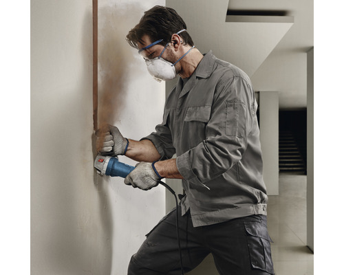 Remeselník pracuje s drážkovacou frézou na stene. Má ochranné okuliare, rúško a rukavice.