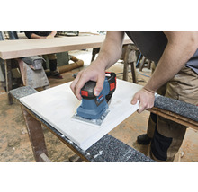 Osoba brúsi drevenú dosku elektrickou vibračnou brúskou na pracovnom stole.