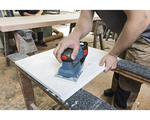 Osoba brúsi drevenú dosku elektrickou vibračnou brúskou na pracovnom stole.