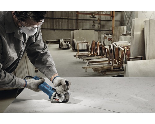 Pracovník s ochranou očí a úst používa uhlovú brúsku na spracovanie kamennej dosky v dielni.