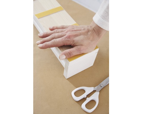 Ruka pevne tlačí na drevenú lištu s lepiacimi pásikmi, nožnice sú pripravené.