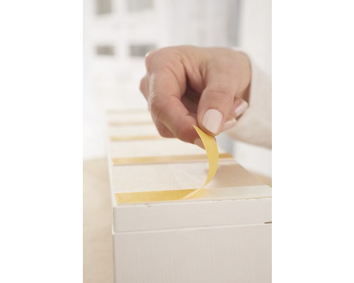 Ruka odstraňuje ochrannú fóliu z lepiacej pásky na bielom predmete.