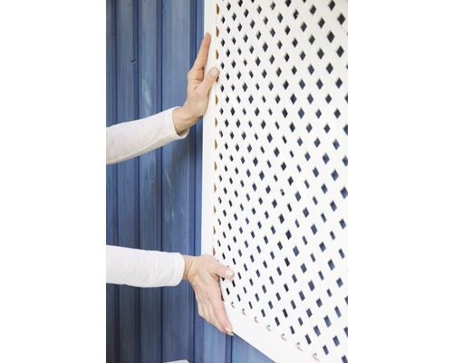 Osoba montuje biely lamelový plotový prvok na modrú drevenú stenu