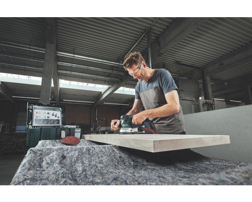 Muž brúsi drevenú dosku excentrickou brúskou v dielni.