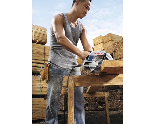 Muž reže drevenú dosku kotúčovou pílou, v pozadí hromady dreva.