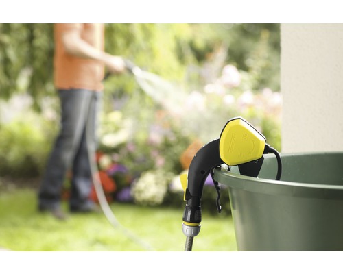 Záhradná scéna so zavlažovacou tyčou pripevnenou k vedru a osobou v pozadí zalievajúcou záhradu.
