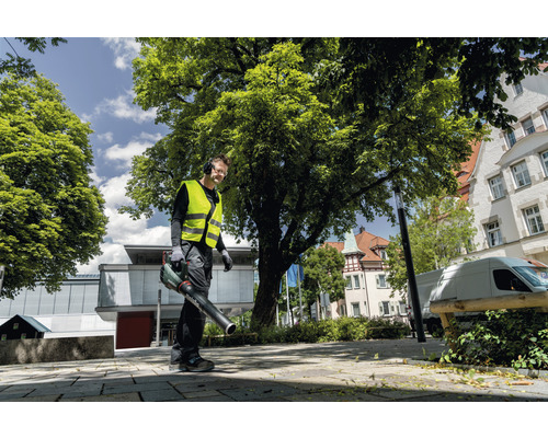 Muž s bezpečnostnou vestou a slúchadlami používa vonku fúkač lístia.