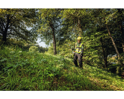 Osoba s ochrannou prilbou a bezpečnostnou vestou pracuje s krovinorezom v zalesnenej oblasti.