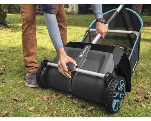 Osoba obsluhuje zberač trávy Gardena na trávniku s lístím.