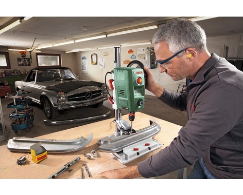 Dielňa s vŕtačkou Bosch, muž vŕta do obrobku a má ochranné okuliare a chrániče sluchu.