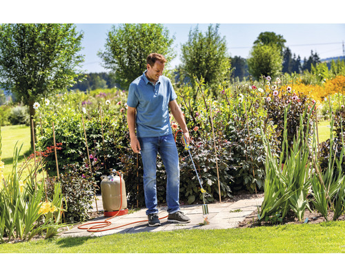 Muž odstraňuje burinu plynovým horákom na kamennej terase v záhrade.