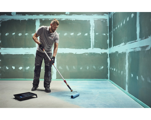 Muž natiera podlahu valčekom v nedokončenej miestnosti.