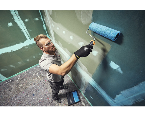 Muž maľuje valčekom stenu v miestnosti so sadrokartónovými stenami a ochrannou rúnnou podlahou.