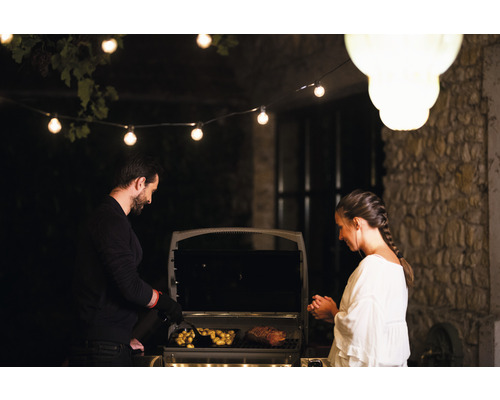 Muž a žena grilujú mäso a zemiaky na vonkajšom grile osvetlenom svetelnou reťazou.