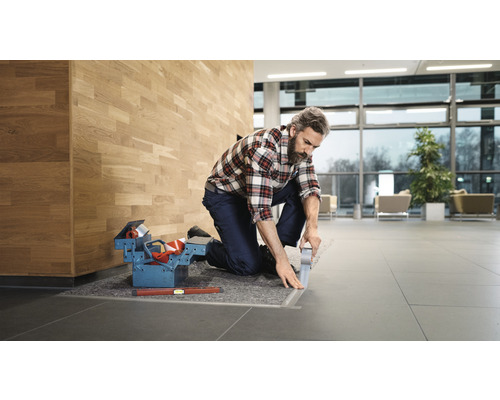 Remeselník kľačí a prilepuje podlahovú lištu v budove. Debnička s náradím a vodováha sú pripravené.