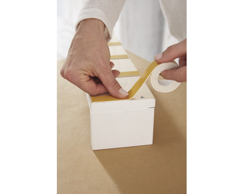 Osoba pripevňuje obojstrannú lepiacu pásku na bielu drevenú krabicu.