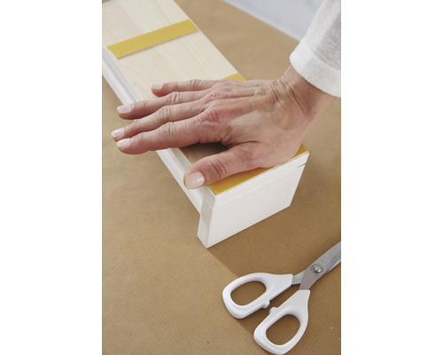 Ruka pritláča drevenú lištu s lepiacou páskou na podklad, vedľa ležia nožnice.