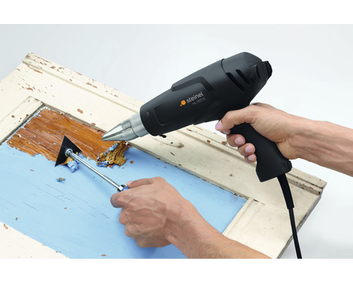 Osoba odstraňuje farbu z okenného rámu pomocou teplovzdušnej pištole a škrabky na farbu