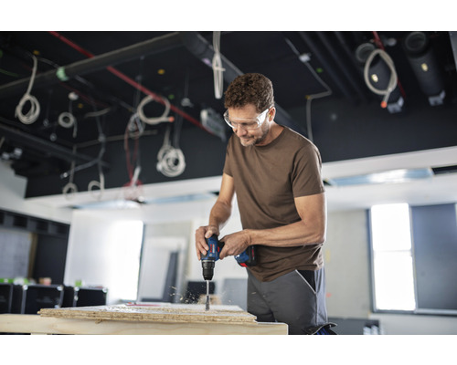 Muž vŕta vŕtačkou do drevenej dosky. Má ochranné okuliare.