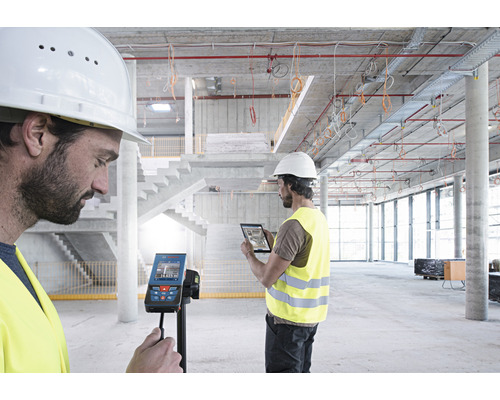 Dvaja stavební robotníci používajú digitálny laserový diaľkomer a tablet na stavenisku.