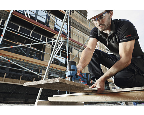 Stavebný robotník s ochrannou helmou a okuliarmi reže drevené dosky priamočiarou pílou na stavenisku. V pozadí je lešenie.