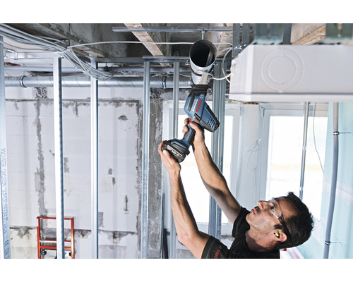 Remeselník montuje potrubie k stropu v hrubej stavbe pomocou akumulátorového skrutkovača.