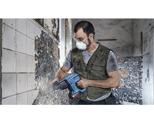 Pracovník s ochrannými okuliarmi, maskou a ochranou sluchu používa vŕtacie kladivo na stenu.