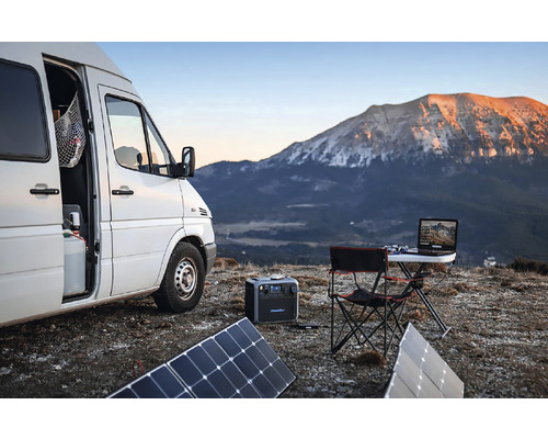 Scéna s dodávkou, solárnymi panelmi, prenosnou elektrárňou, skladacím stolom a notebookom na pozadí hory