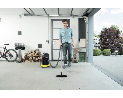Muž používa viacúčelový vysávač v garáži na odstránenie nečistôt. V pozadí je vidieť gril, drevo a bicykel.
