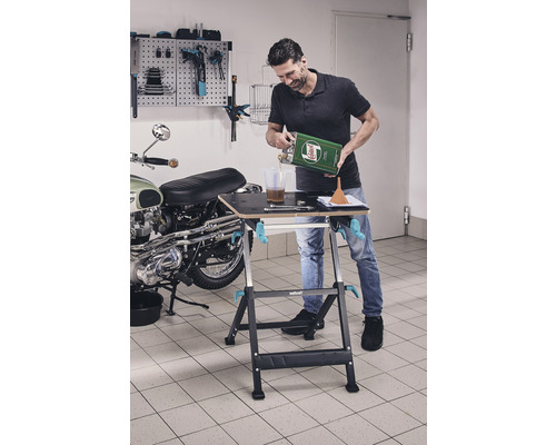 Muž prelieva olej na pracovnom stole v garáži, v pozadí motocykel a stena s náradím.