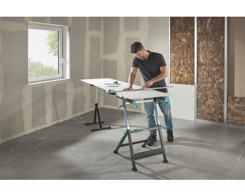 Muž reže sadrokartónovú dosku na pracovnom stole Wolfcraft v hrubej stavbe.