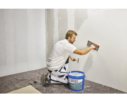 Muž nanáša hladkú stierku na stenu pomocou hladidla. V popredí je vedro hladkej stierky.