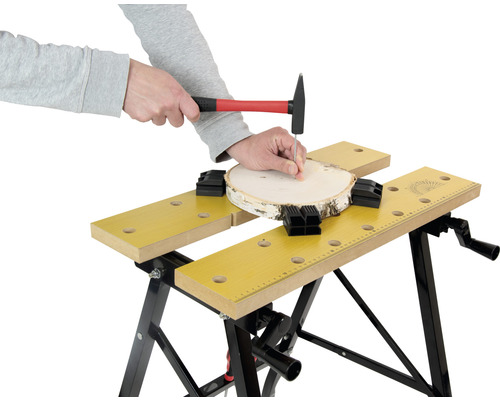 Osoba zatĺka kladivom klinec do kusa dreva, ktorý je upevnený pomocou pracovného stola.