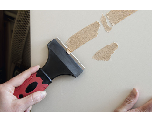 Muž odstraňuje staré zvyšky lepiacej pásky zo steny škrabkou.