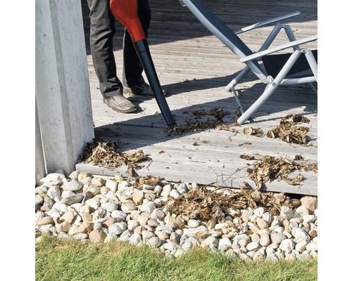 Osoba používa fúkač lístia na odstránenie lístia z drevenej terasy. V pozadí je vidieť ležadlo.