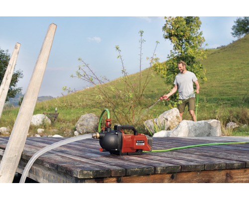 Domáca vodáreň Einhell na drevenom móle s mužom v pozadí, ktorý polieva záhradnou hadicou